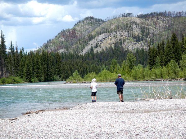 Two people are fishing in a river with mountains in the background.