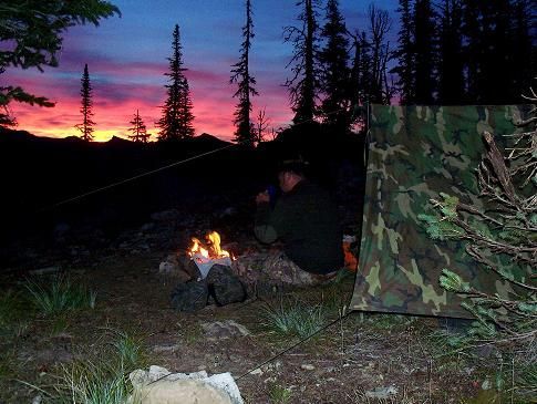 A man is sitting around a campfire in the woods at sunset.