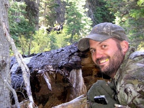 A man in a camo hat is smiling in front of a tree stump.