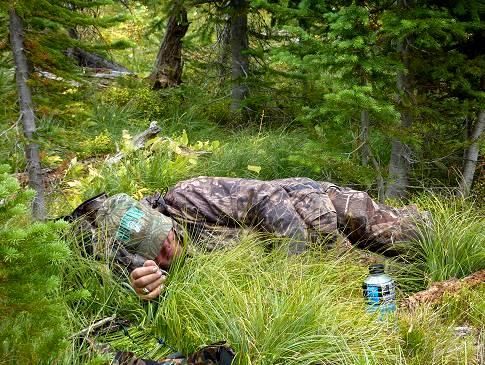 A man in a camouflage suit is laying in the grass in the woods.