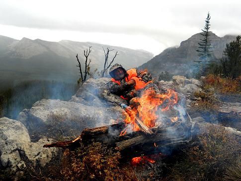 A man is sitting in front of a fire in the mountains.