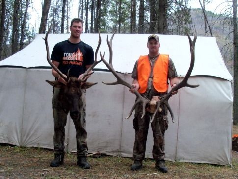 Two men standing in front of a tent holding deer antlers