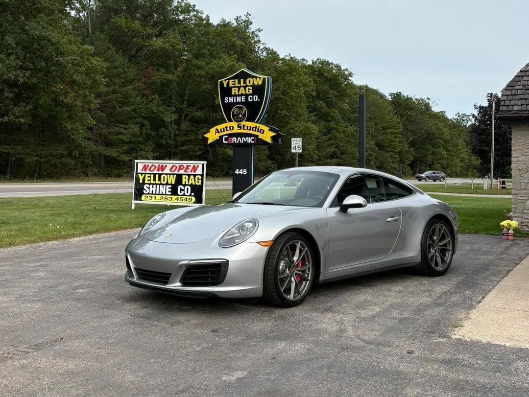 Silver Porsche coupe parked in front of Yellow Hat Shine car wash sign.