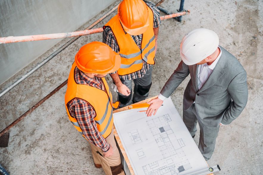 Tre persone con caschi di sicurezza e indumenti da lavoro in piedi su un pavimento di cemento osservano la planimetria di un edificio.