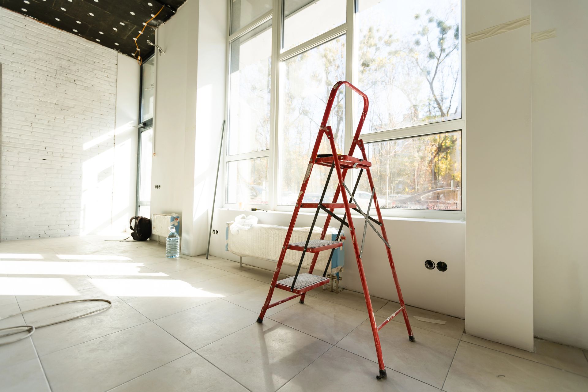 In una stanza luminosa e moderna con ampie finestre e pareti bianche, una scala a pioli rossa è appoggiata su un pavimento piastrellato.