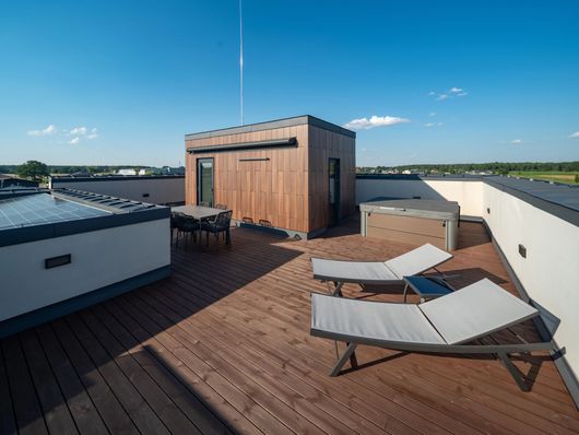 Terrazza panoramica sul tetto con pavimentazione in legno, lettini prendisole, set da pranzo e vasca idromassaggio sotto un cielo azzurro e limpido.