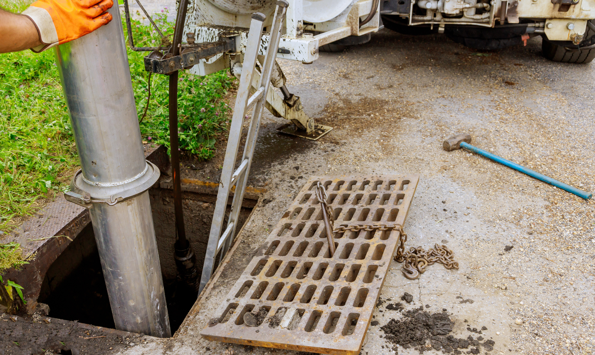 Worker lowering a pipe into a sewer manhole with a truck nearby. Orange-gloved hand is visible.