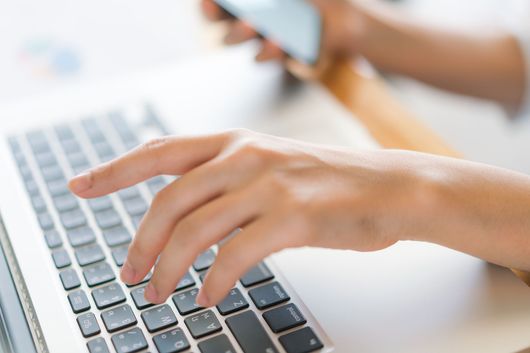 A person is typing on a laptop computer while holding a cell phone.