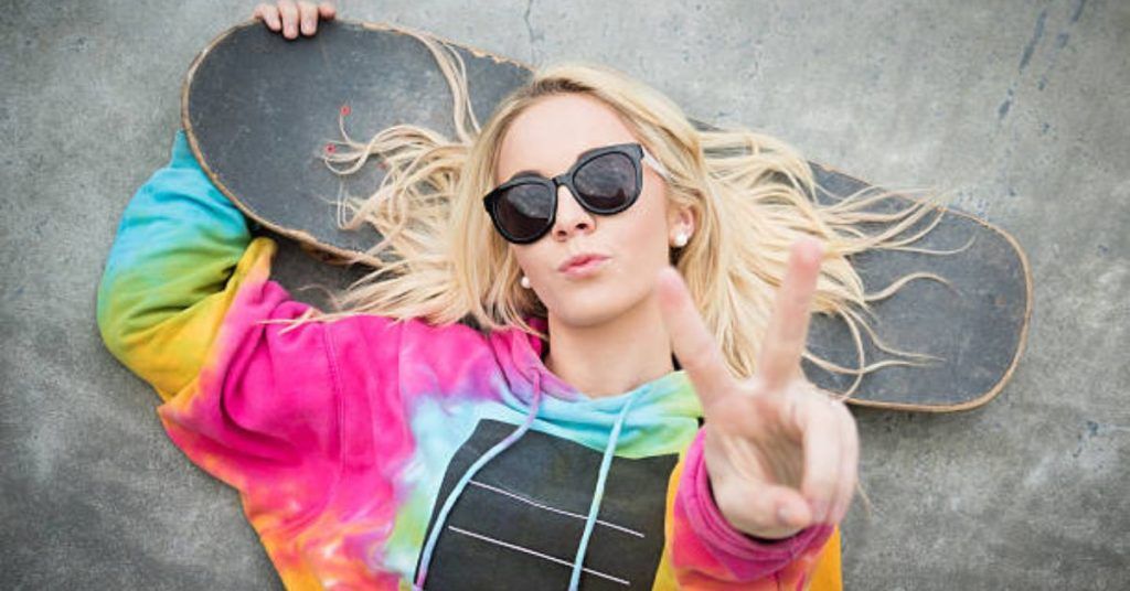 A woman is laying on the ground holding a skateboard and giving a peace sign.