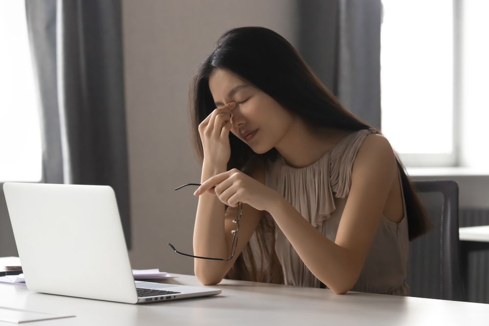 A woman is sitting at a desk with a laptop and rubbing her eyes.