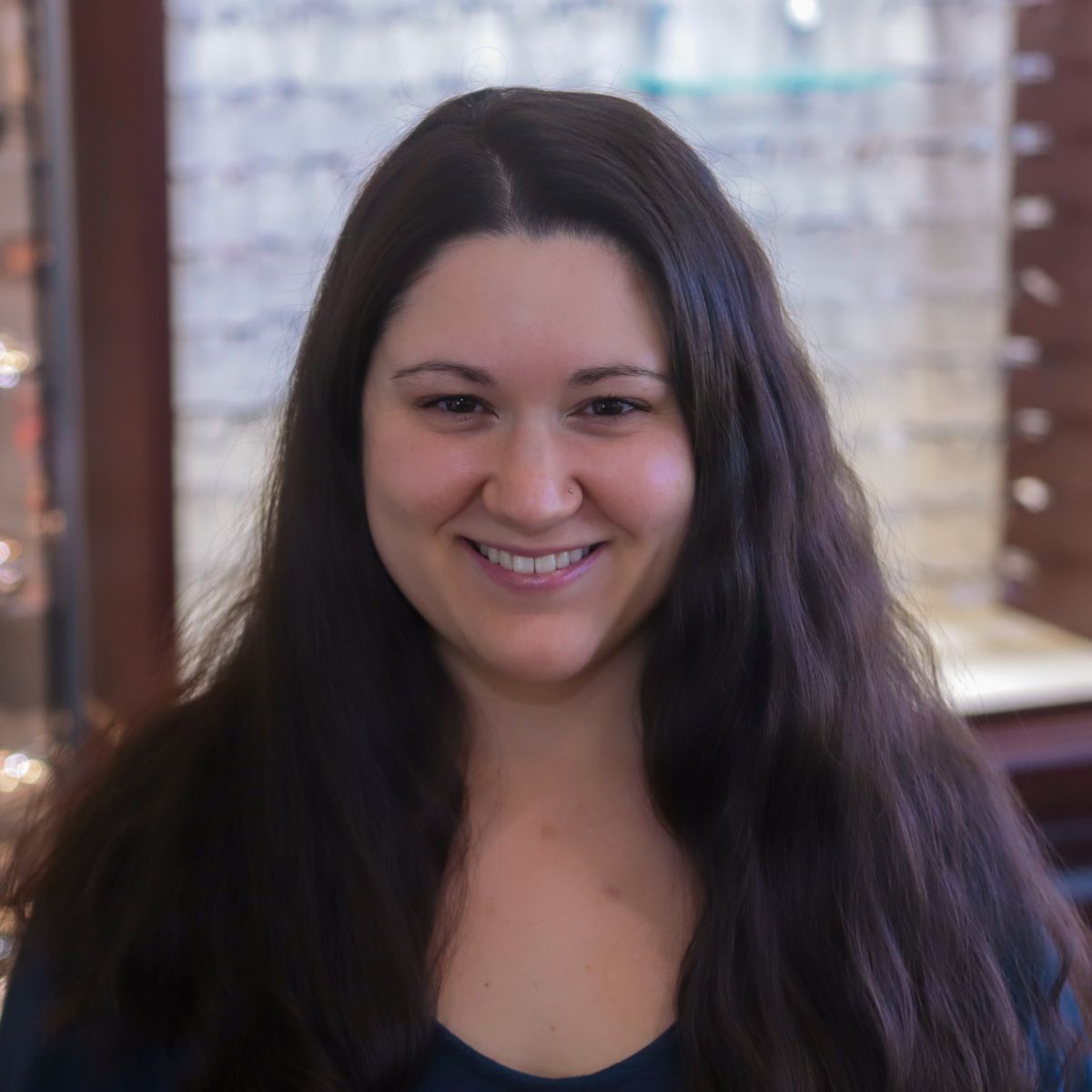 A woman with long dark hair is smiling in front of a display of glasses.