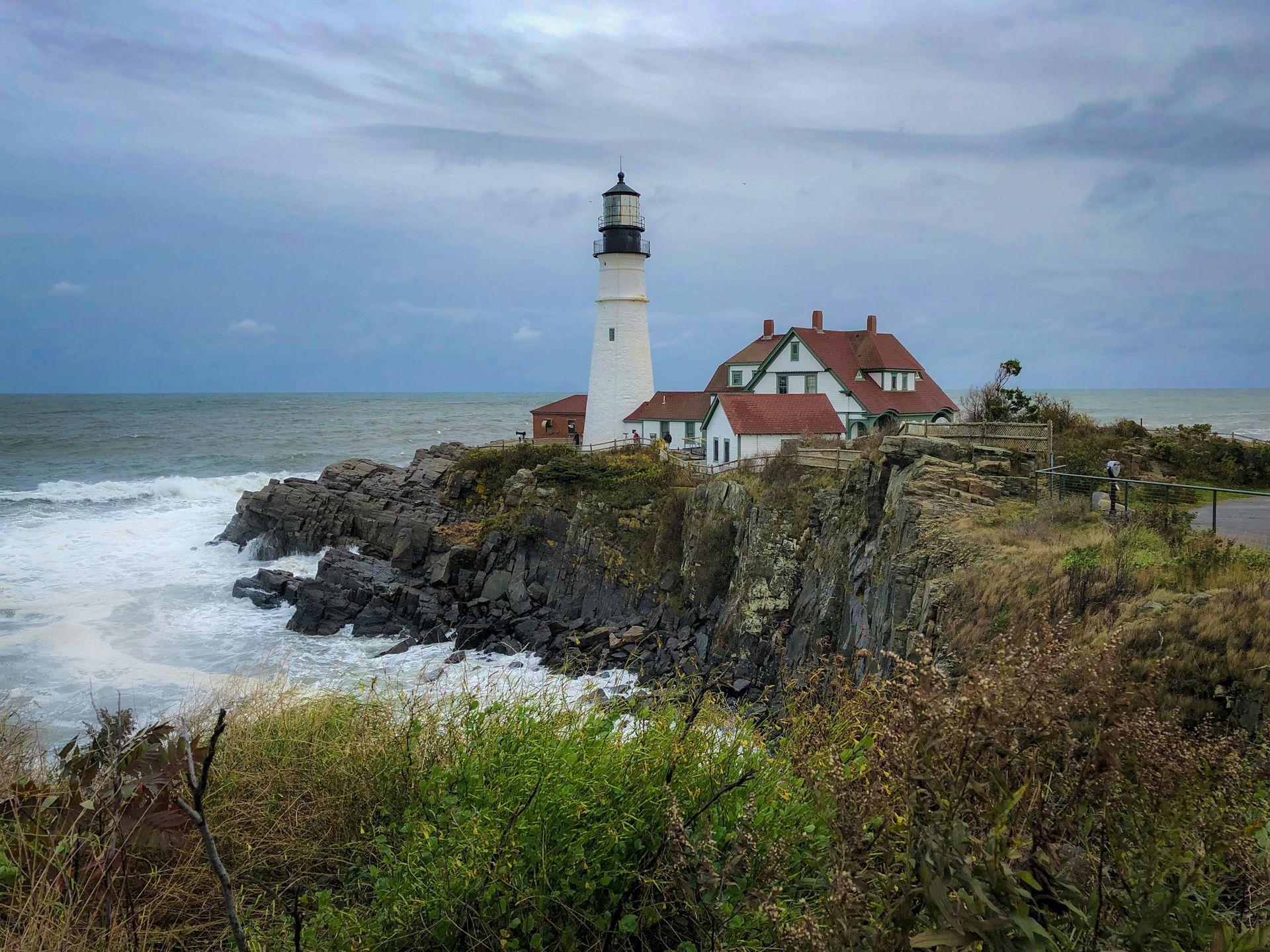 Lighthouse on rocky shore, white tower with attached red-roofed buildings, crashing waves, overcast sky.