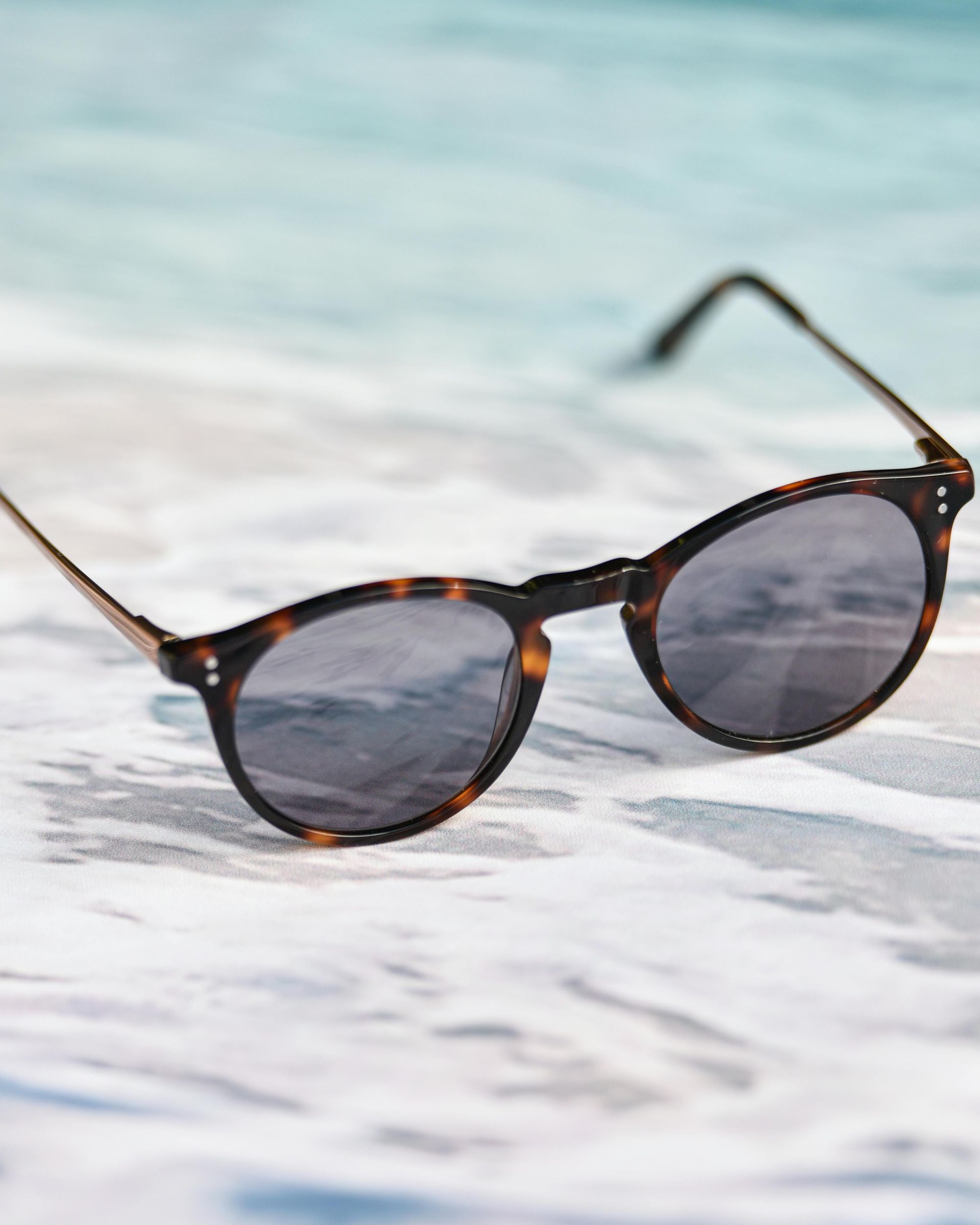 Sunglasses with tortoise frames on a wet sandy surface, with ocean in the background.