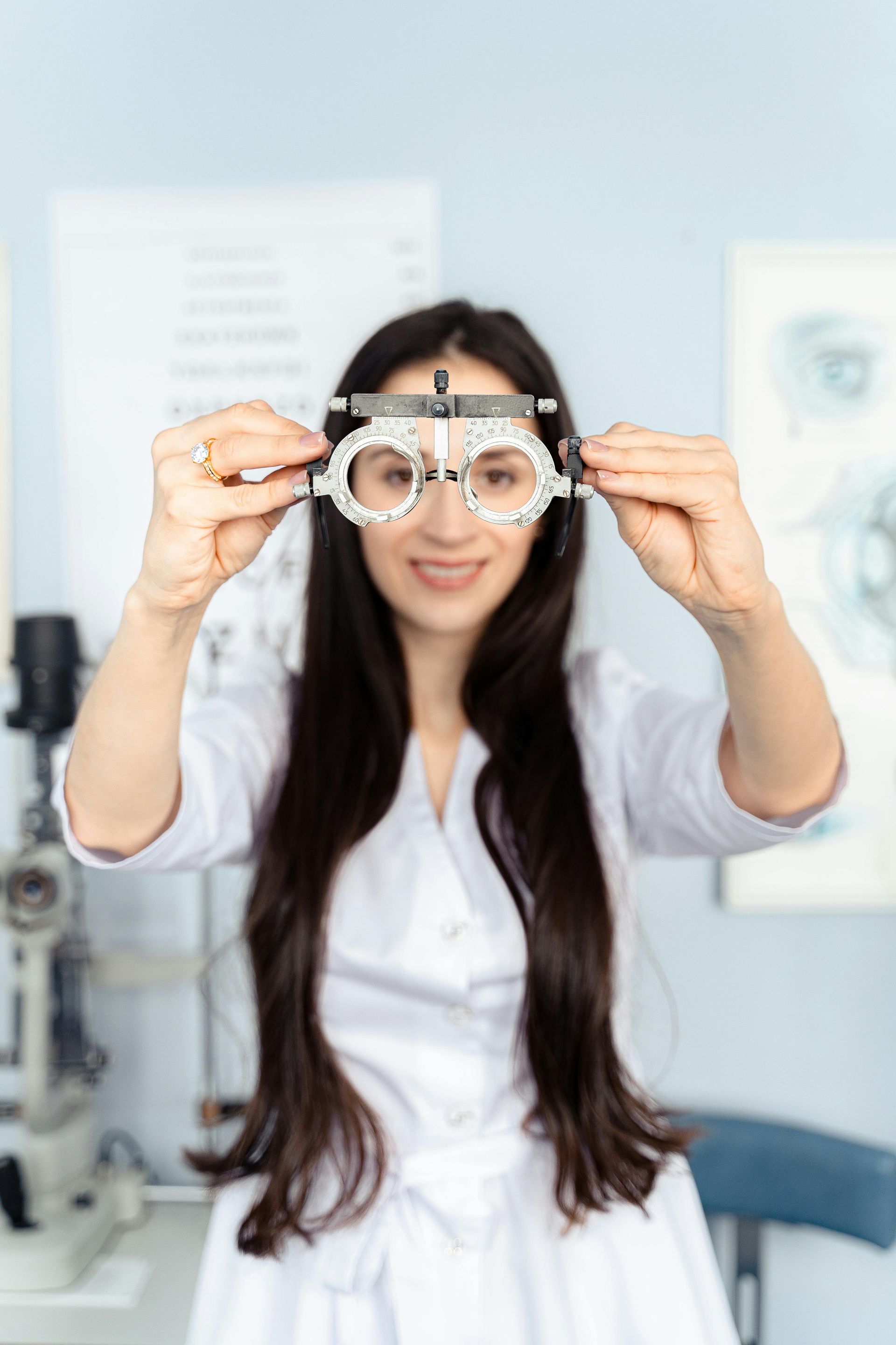 Optometrist holds up a phoropter, smiling, in an exam room.