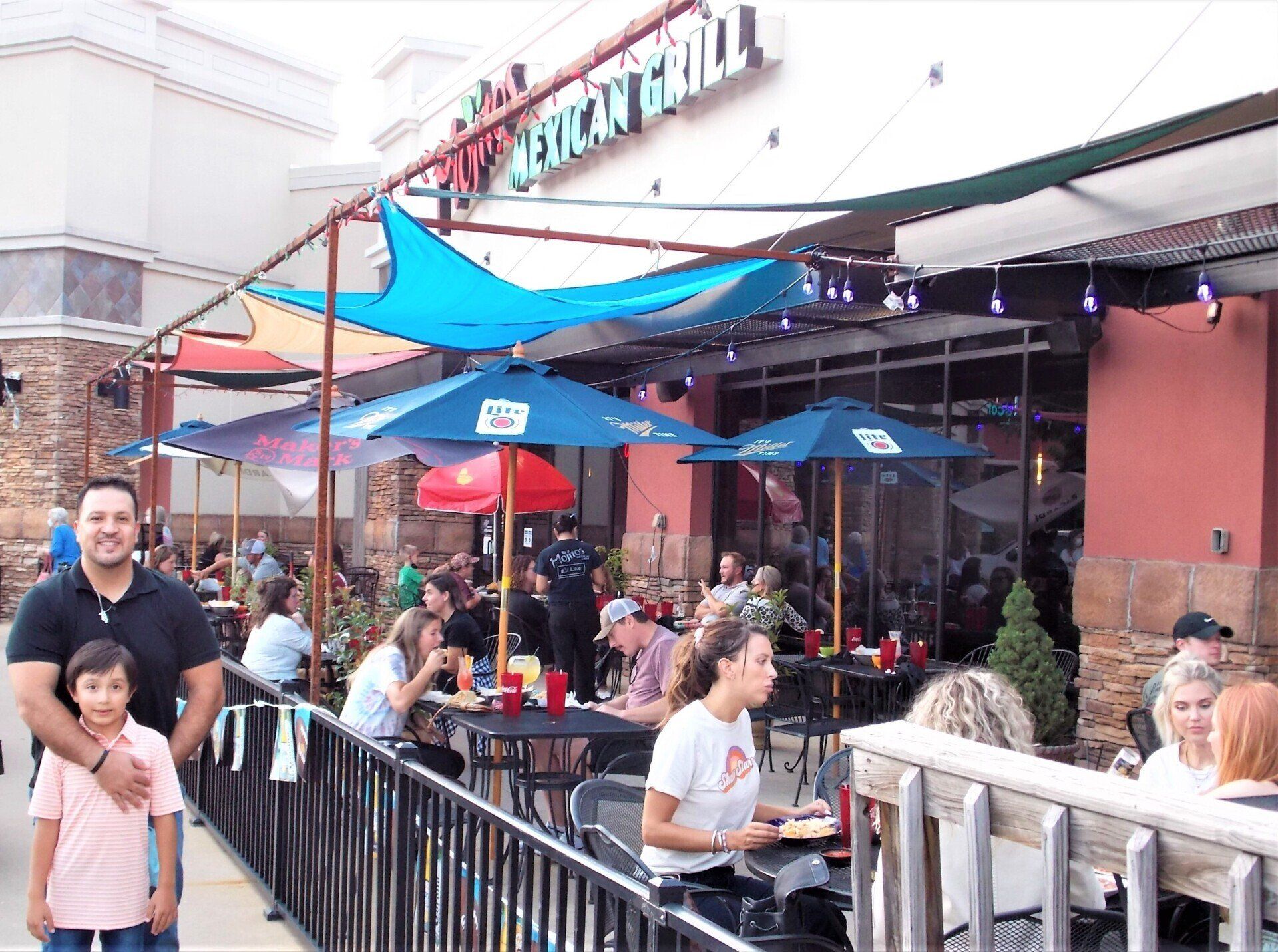 Outdoor patio of Mexican grill, people dining under umbrellas and shade sails. Man and child in foreground.