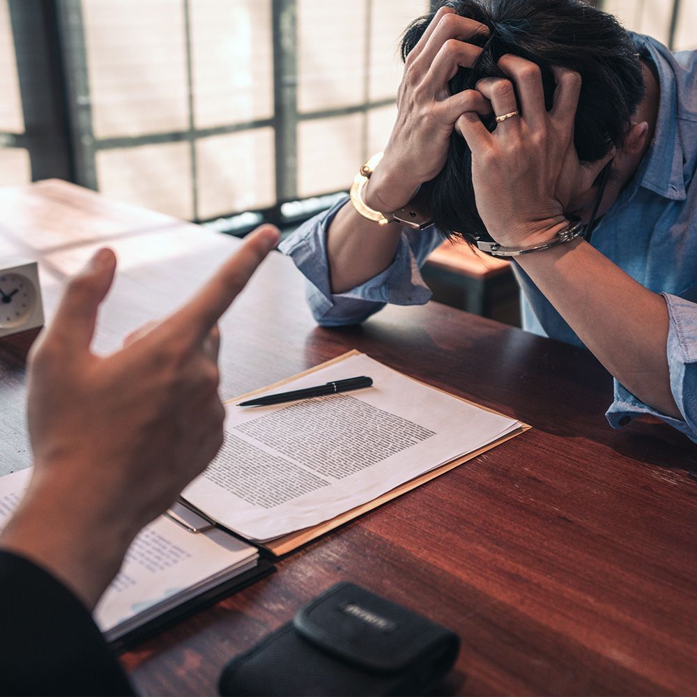 A handcuffed person with hands on head, facing an accusing finger at a table with documents.