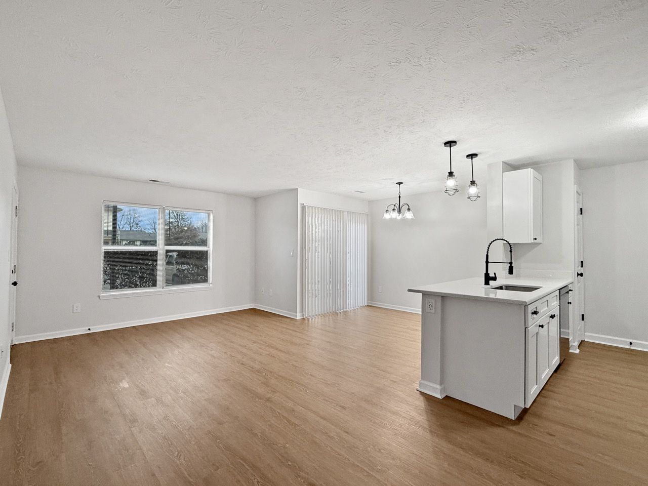 Bright empty living room with wood floors, white walls, pendant lights, window, and a small kitchen island.