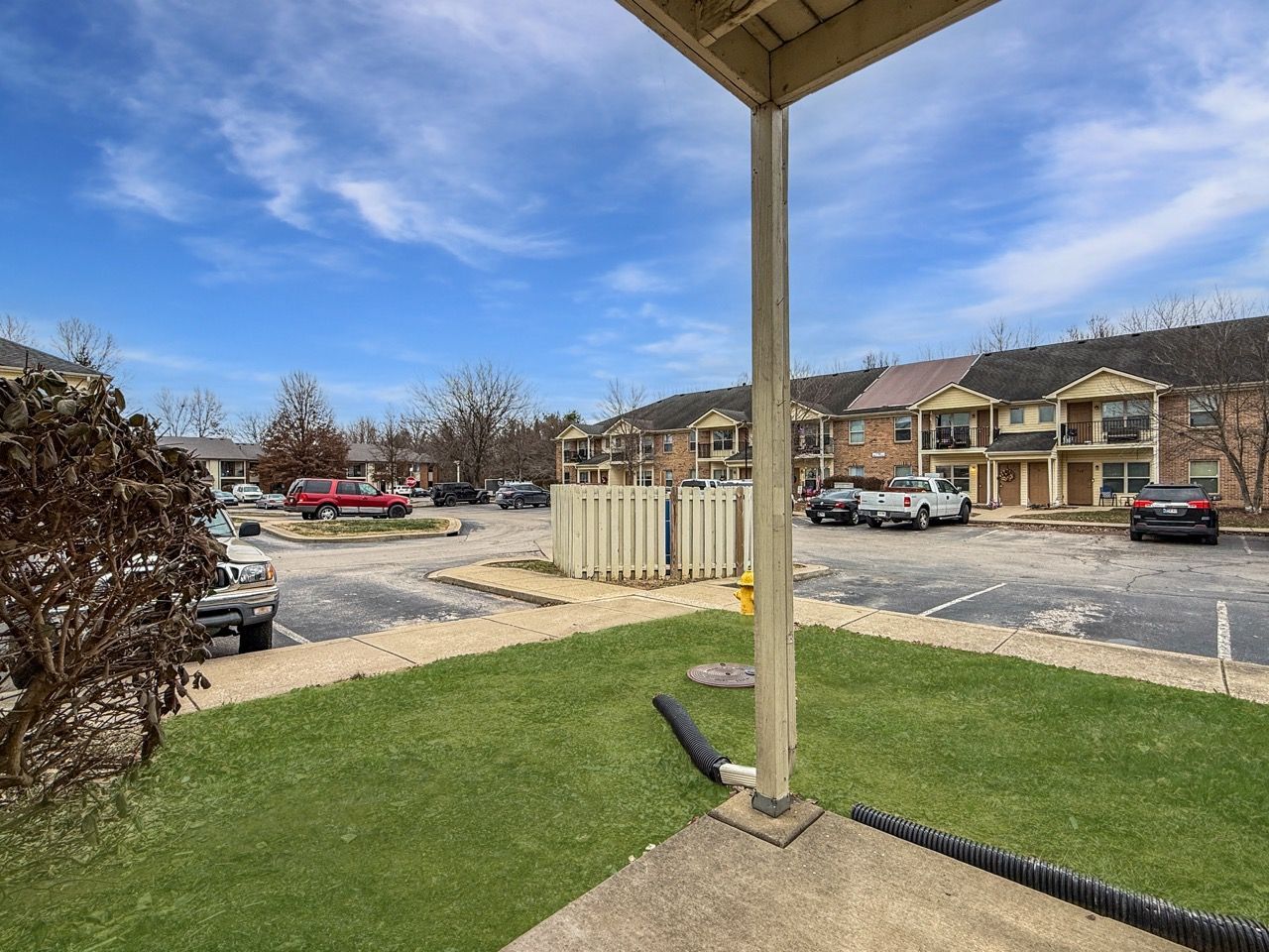 Apartment parking lot with brick buildings, parked cars, and a covered patio post in the foreground