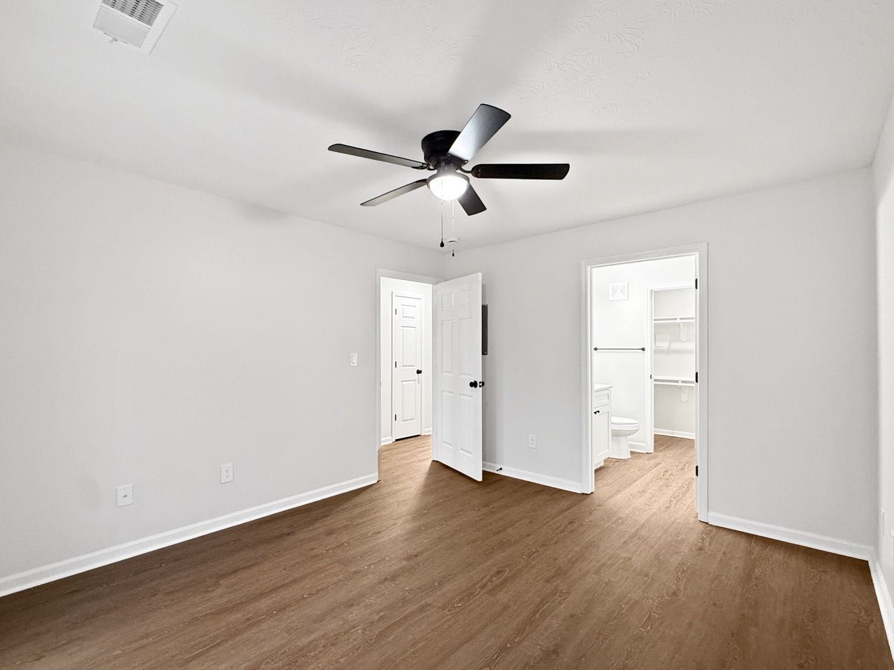 Empty white bedroom with wood floors, ceiling fan, and open doors to a bathroom and hallway