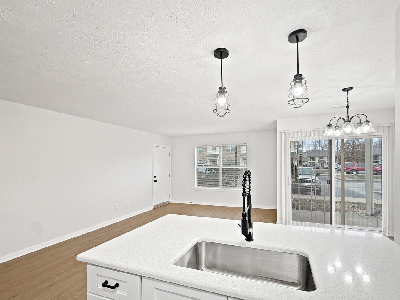 Bright empty kitchen with white walls, wood floor, island sink, pendant lights, and sliding glass doors.