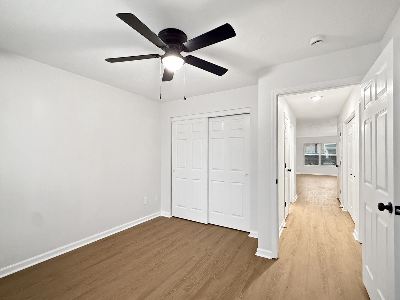 Bright empty bedroom with white walls, wood floors, a ceiling fan, and closet doors, leading to a hallway