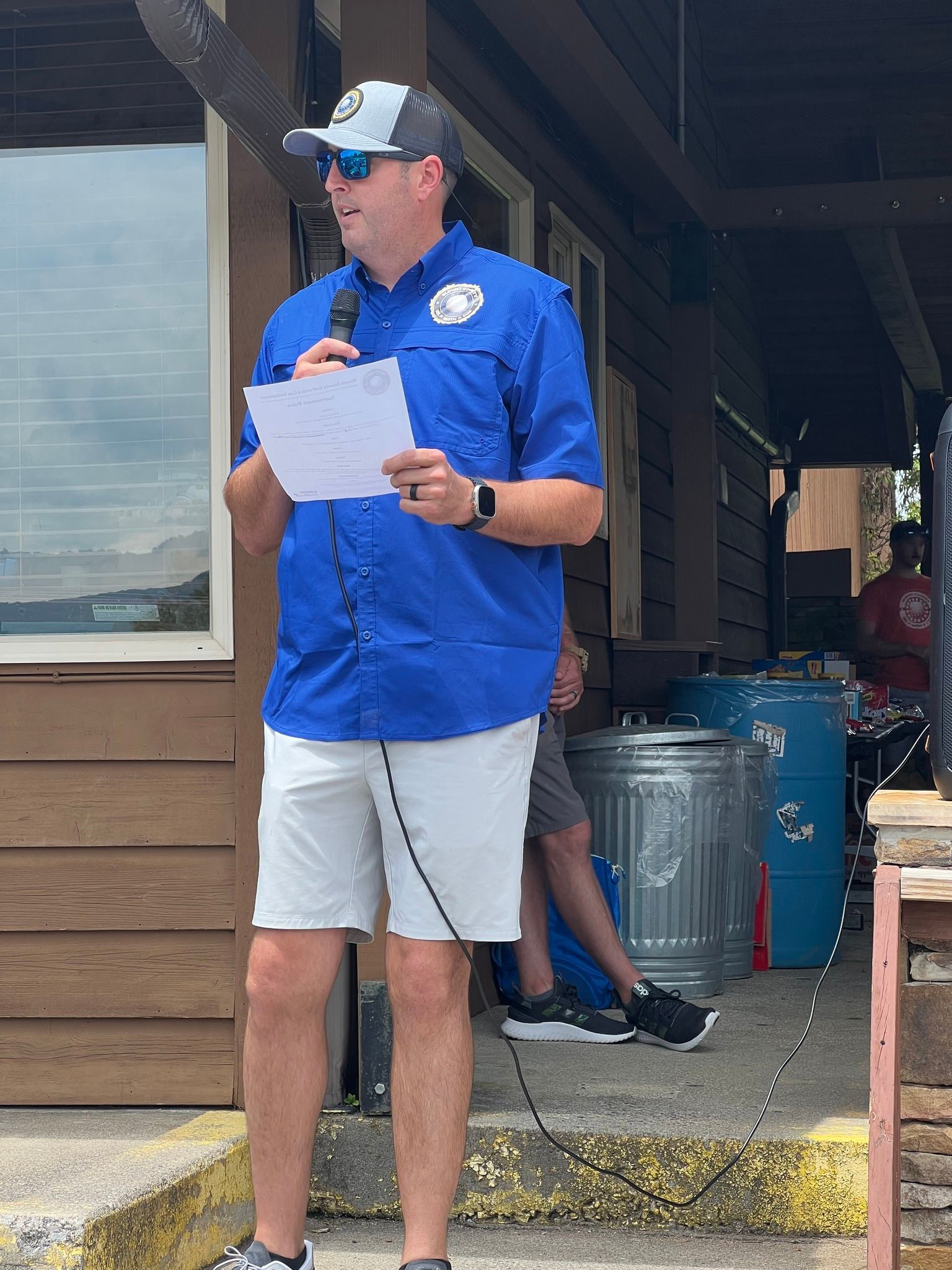 A man is standing in front of a building holding a microphone and a piece of paper.