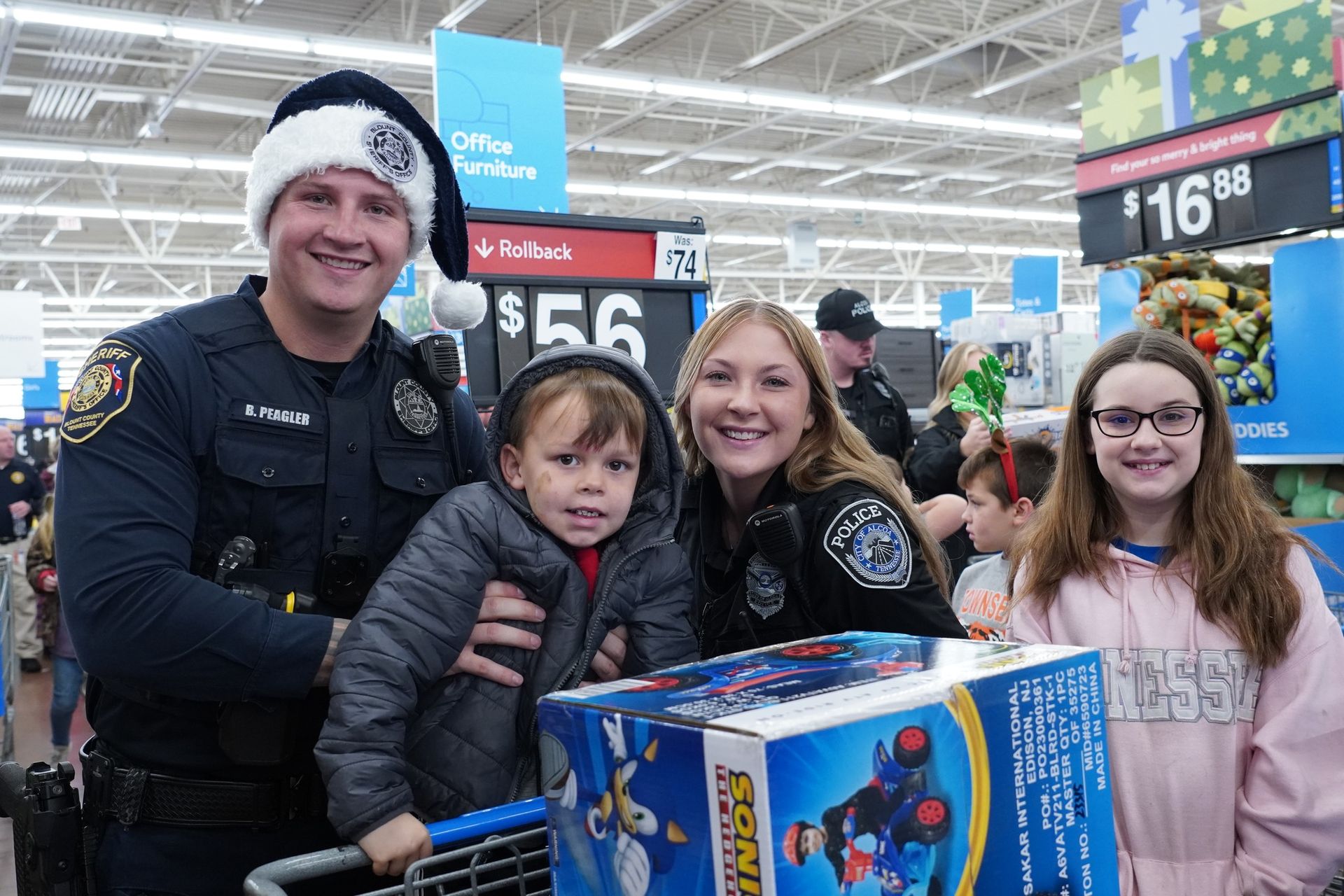 A police officer is holding a child in a shopping cart while a family poses for a picture in a store.