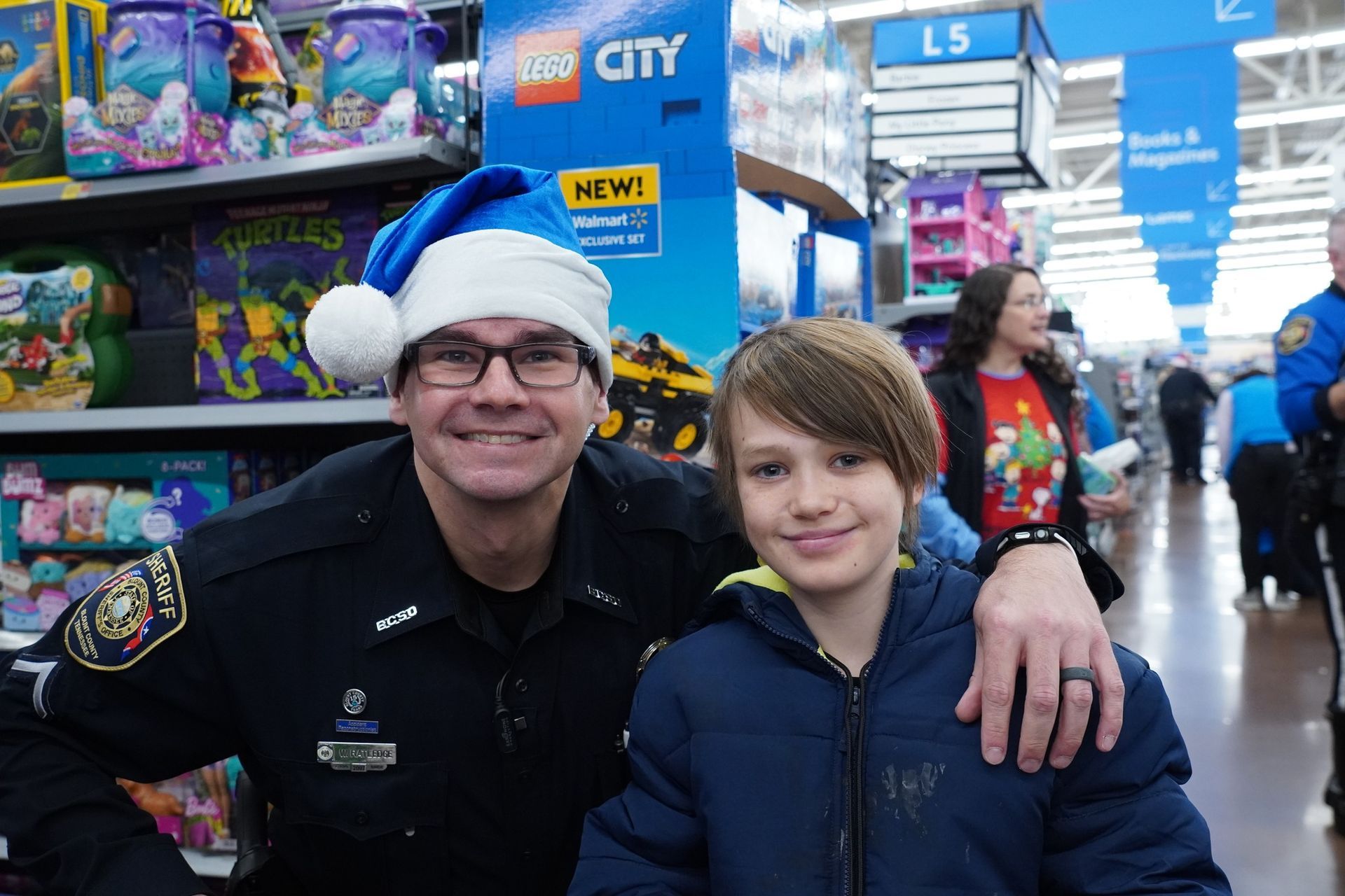 A police officer and a boy are posing for a picture in a store . the boy is wearing a santa hat.