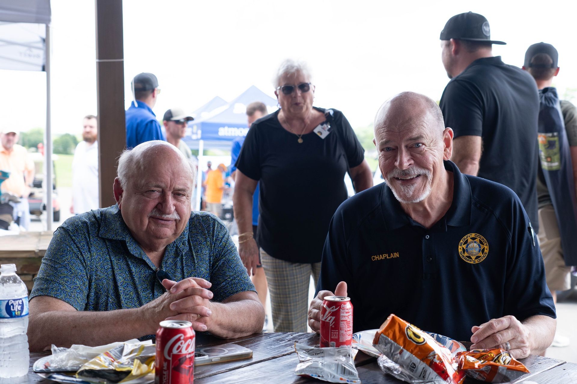 Two men are sitting at a table eating chips and drinking coca cola.