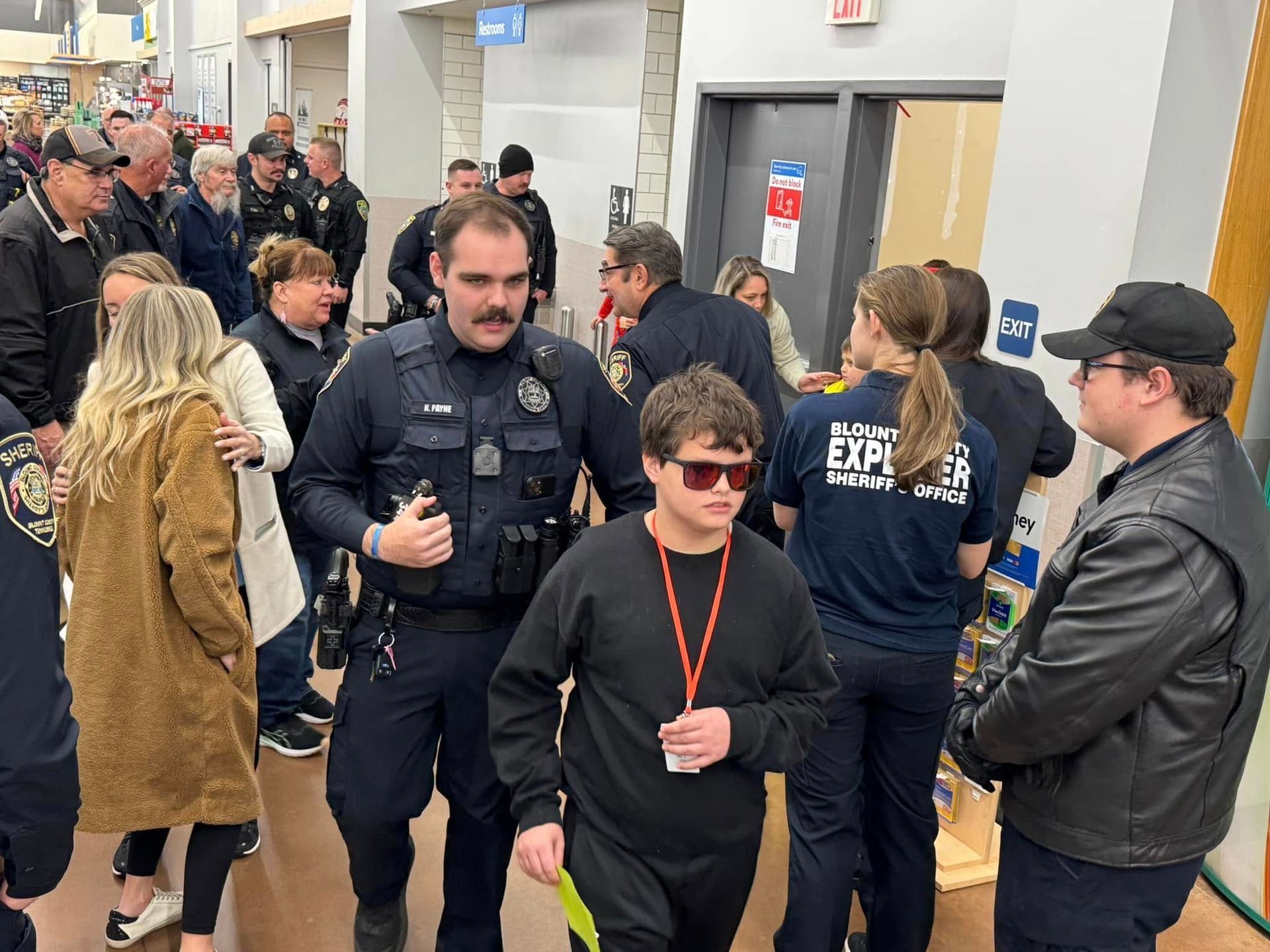 A group of people are standing in a room with a police officer and a boy wearing sunglasses.
