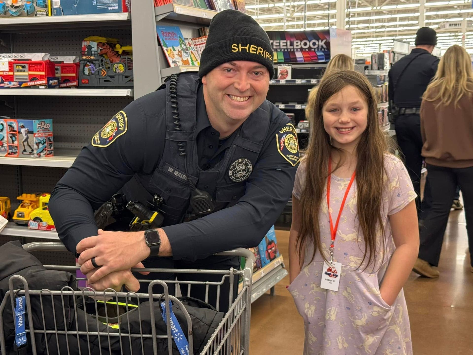 A police officer is standing next to a little girl in a shopping cart.