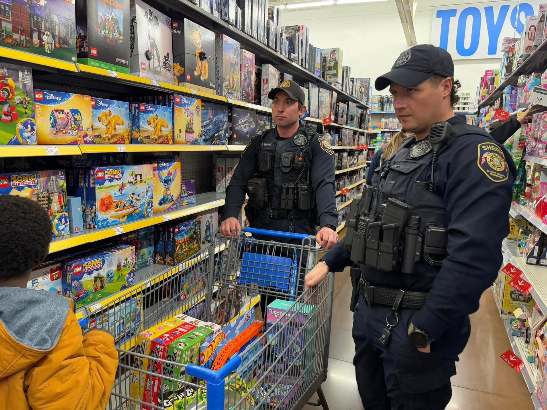 Two police officers are looking at a child in a shopping cart in a toy store.