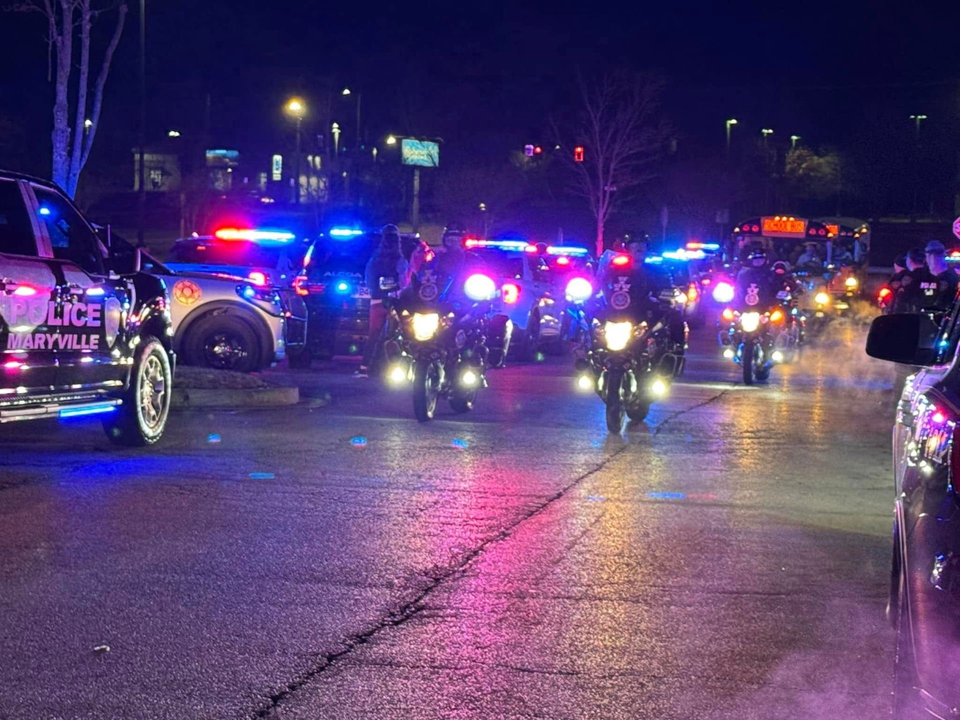 A row of police cars and motorcycles are driving down a street at night.