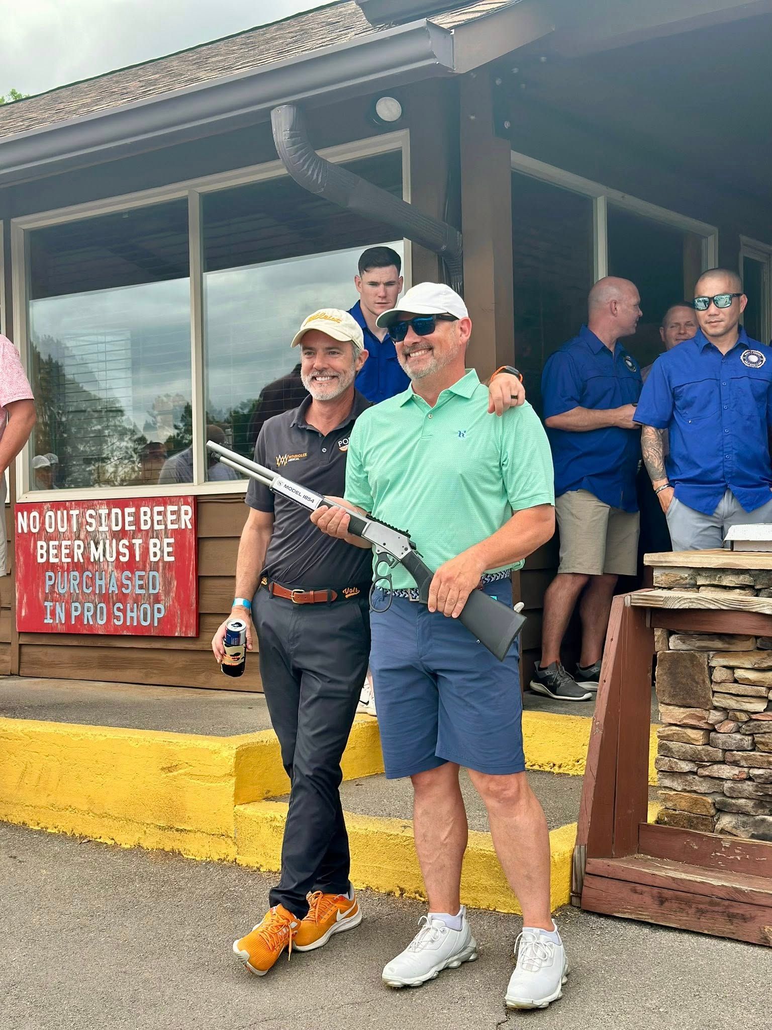Two men posing outside a building, one holding a shotgun. Others in background.