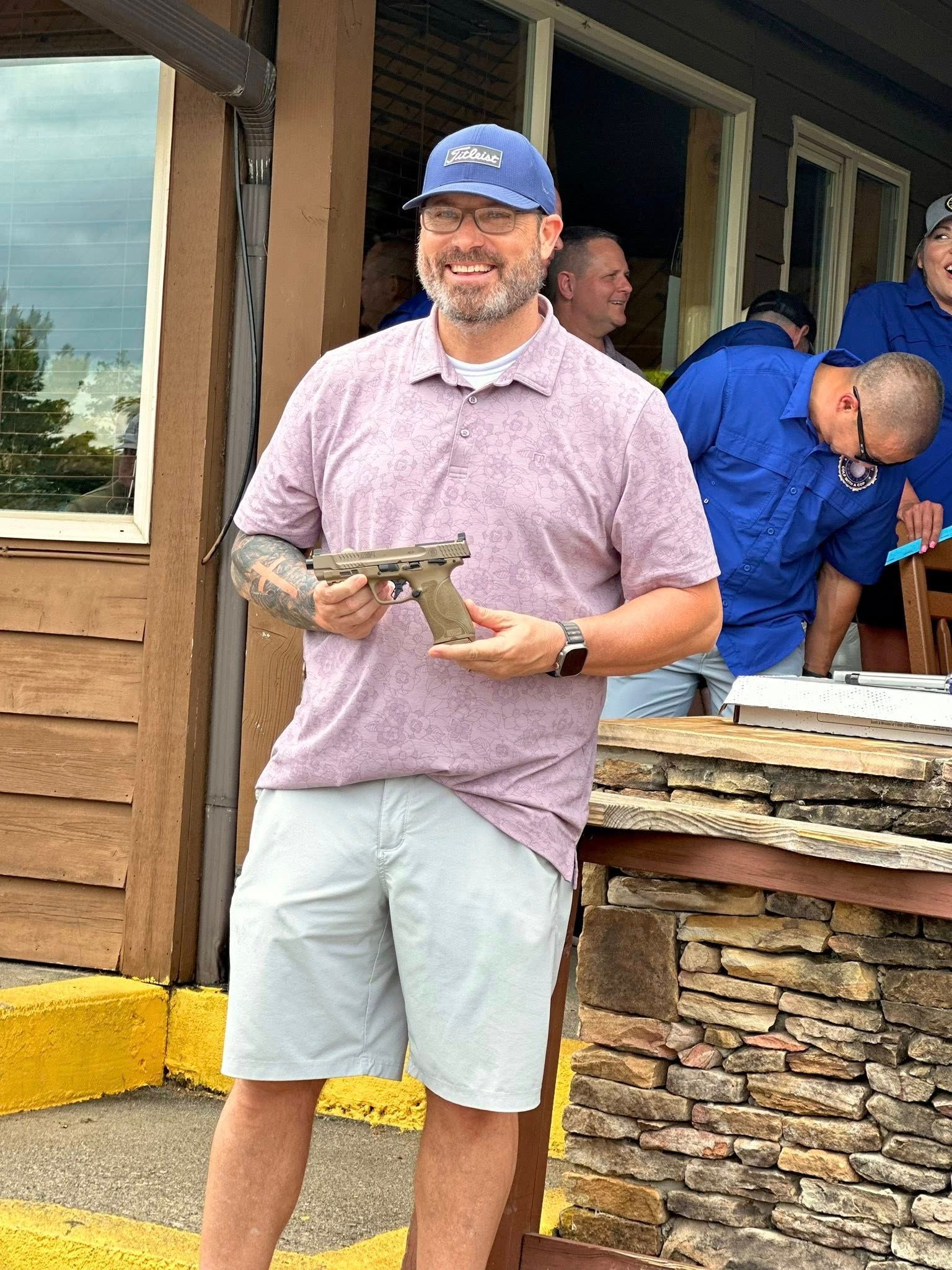 Man in pink shirt and shorts holds a handgun outdoors, others in blue shirts in background.