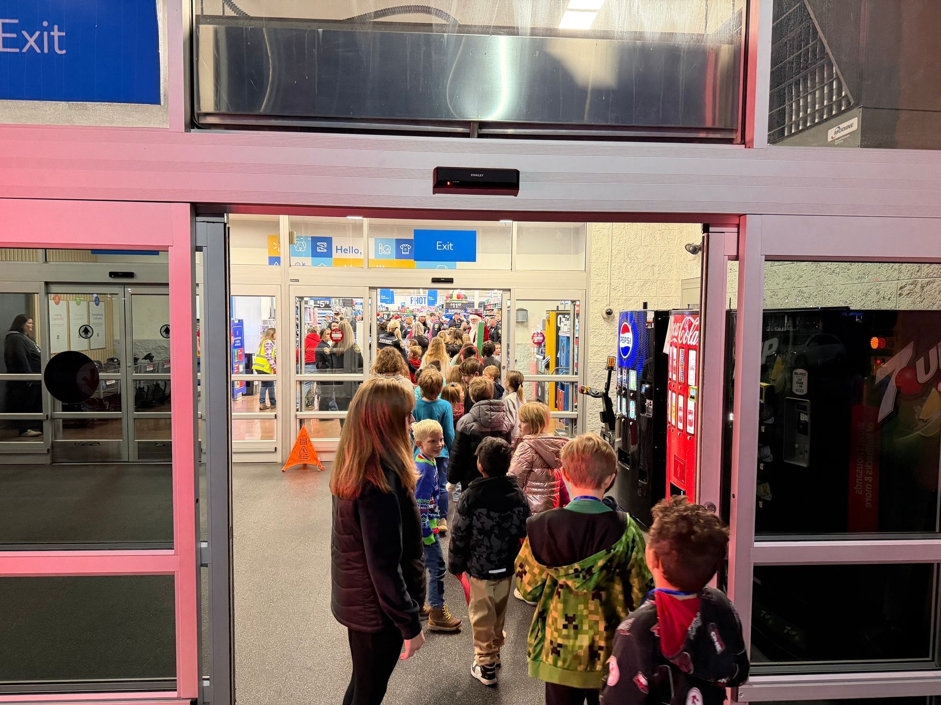 Children lined up entering a store, likely a Walmart, through automatic doors.