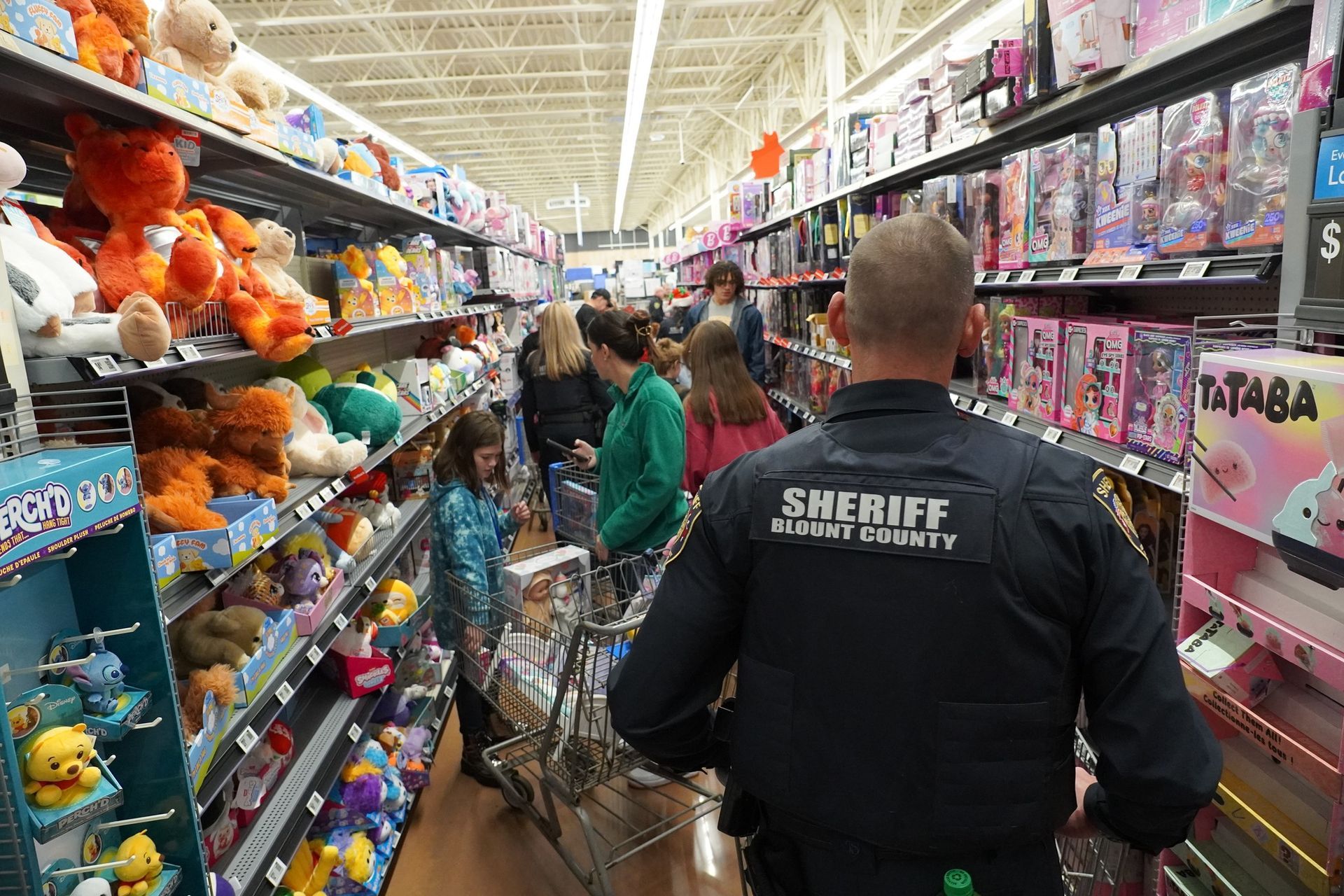 Sheriff in uniform walking through a toy aisle at a store. People shopping, stuffed animals and toys on shelves.