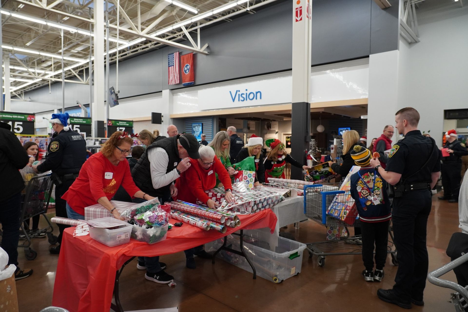 Volunteers wrapping gifts at a table in a store, with shoppers and a police officer nearby.