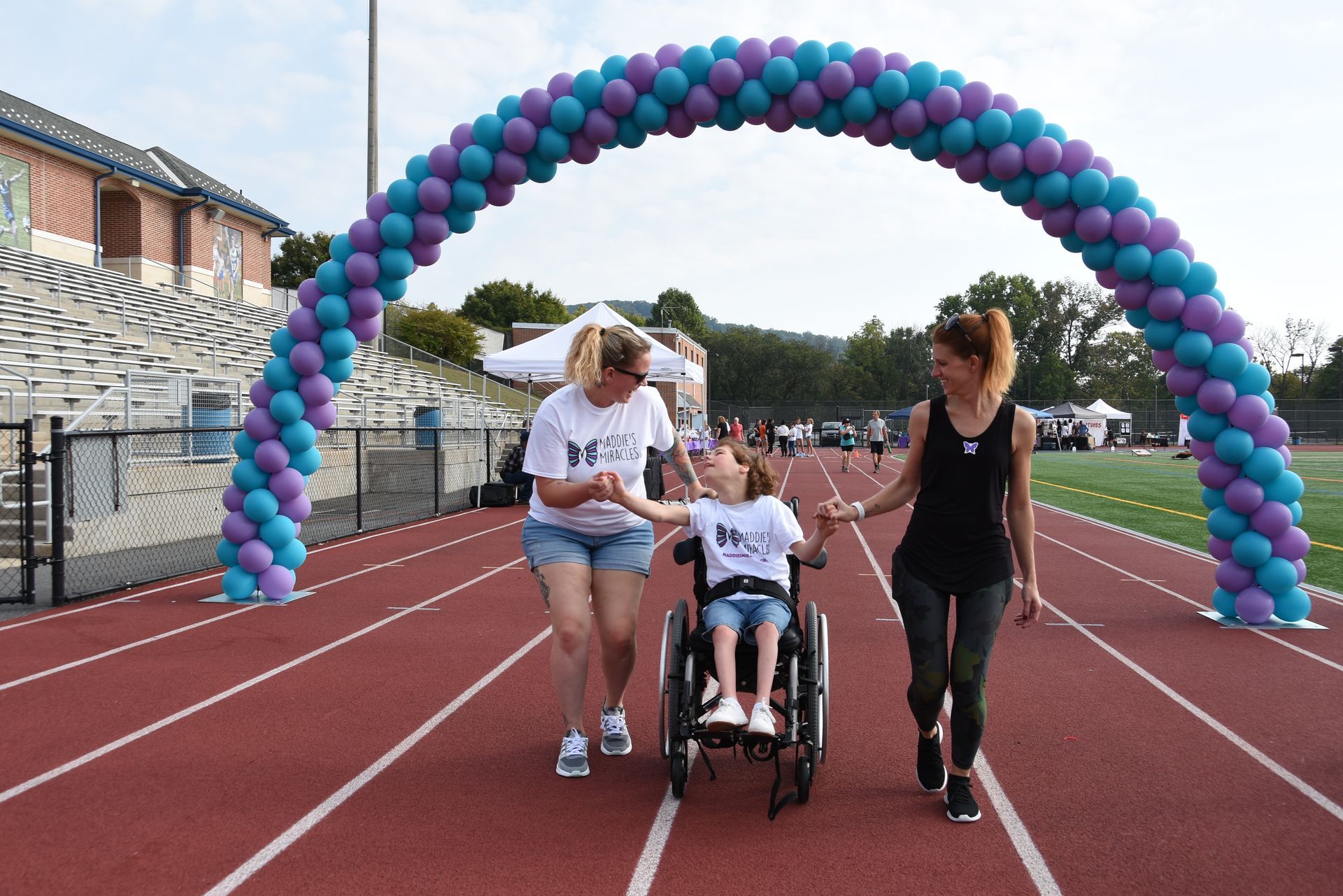 Girl in wheelchair, supported by two women, passes under a balloon arch on a track.