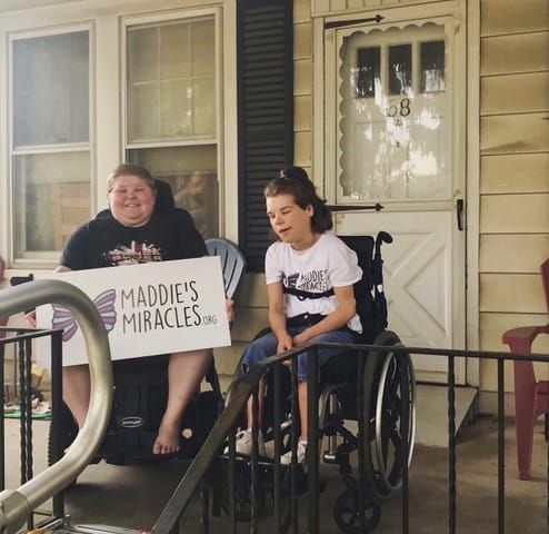 two children sitting on a porch in wheelchairs holding a Maddie's Miracles sign and smiling