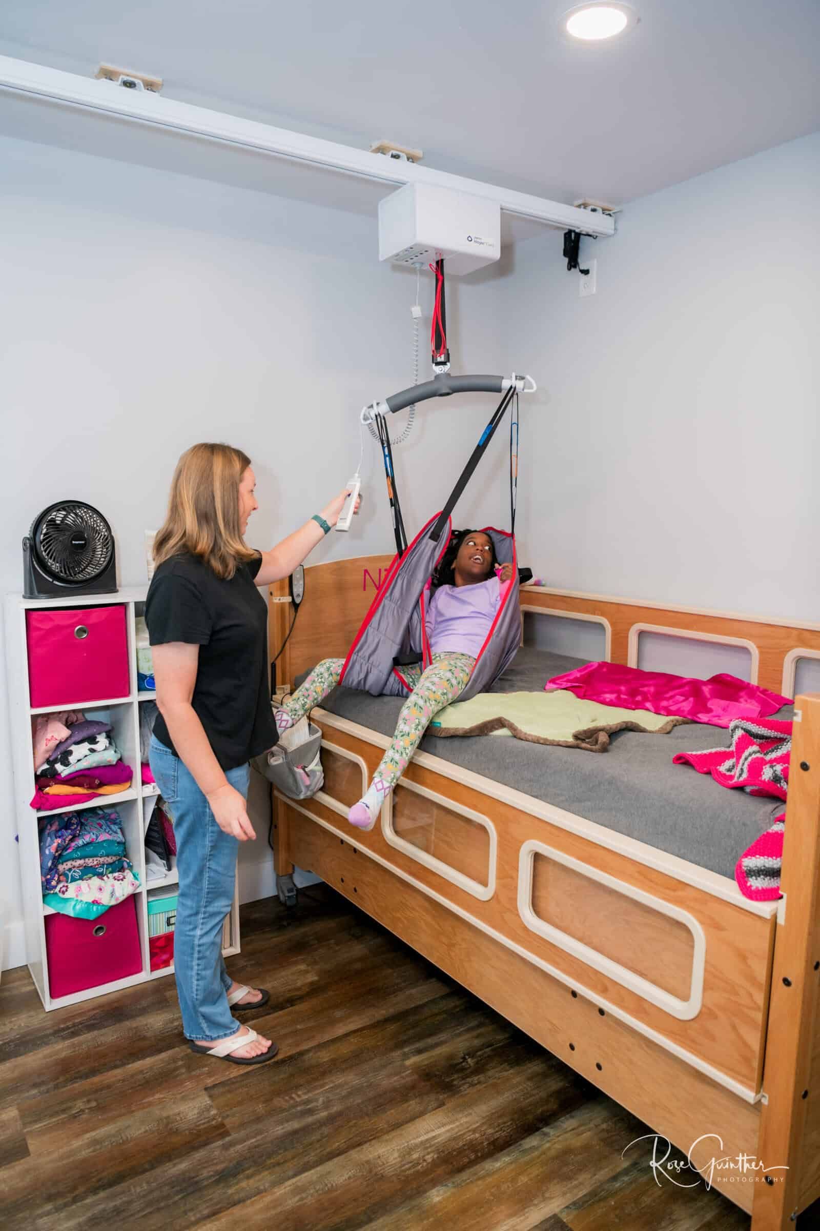 Woman operating ceiling lift, transferring child from bed. White room, wooden bed, pink storage cubes.