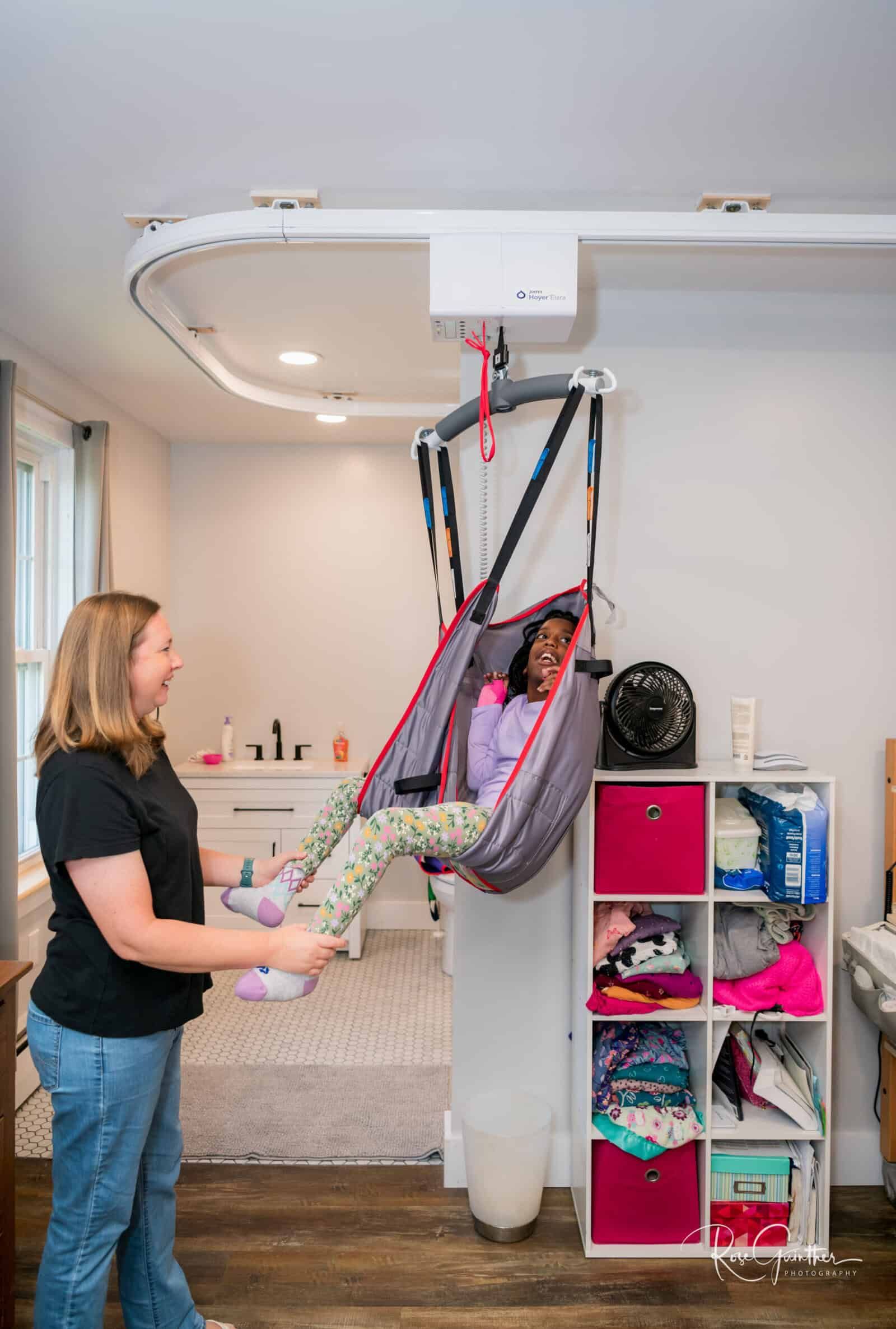 Woman assists girl in ceiling track lift system in a room. The girl smiles.