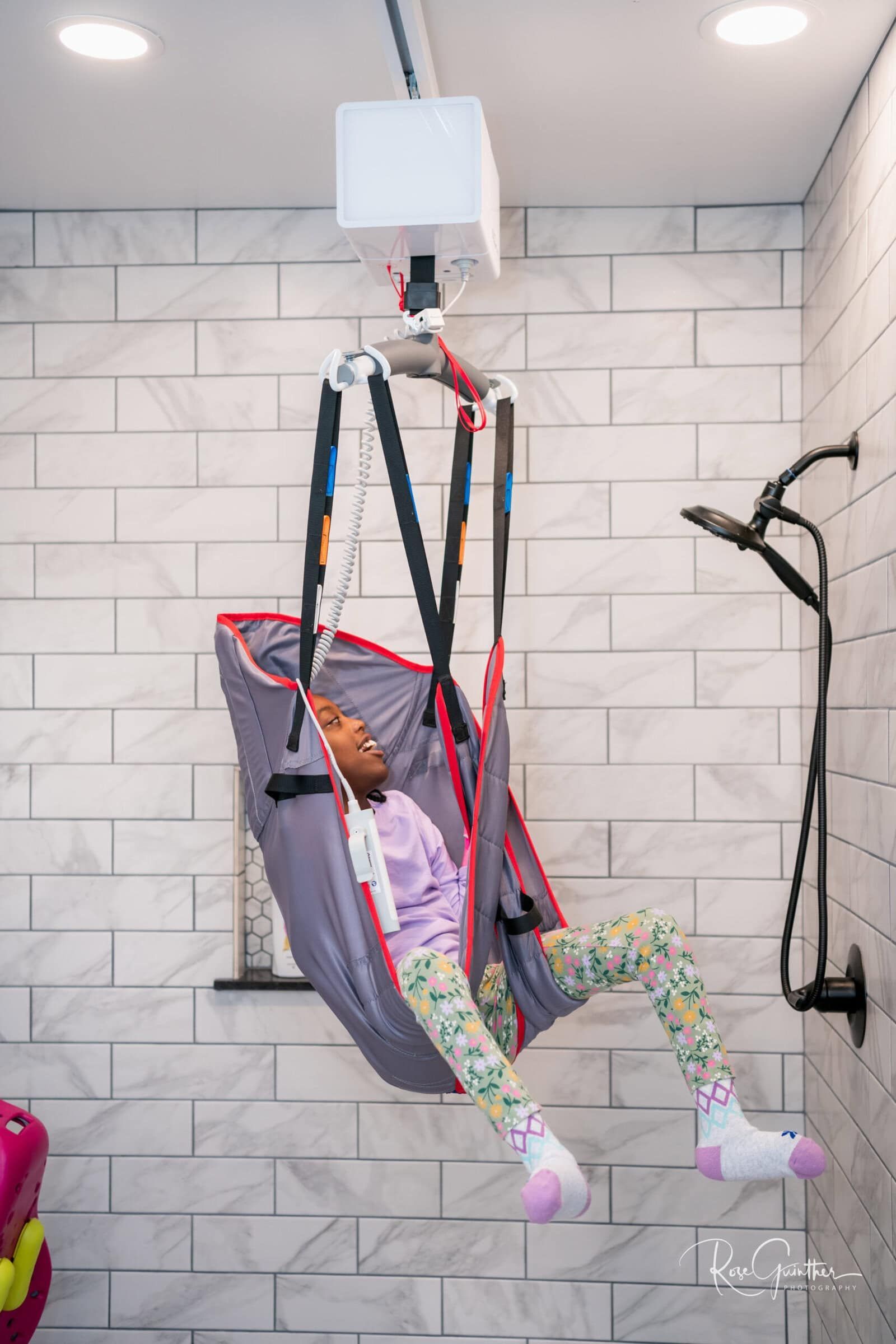 A person in a sling lift in a shower. Light-skinned child in a grey sling, colorful leggings, and pink slippers.