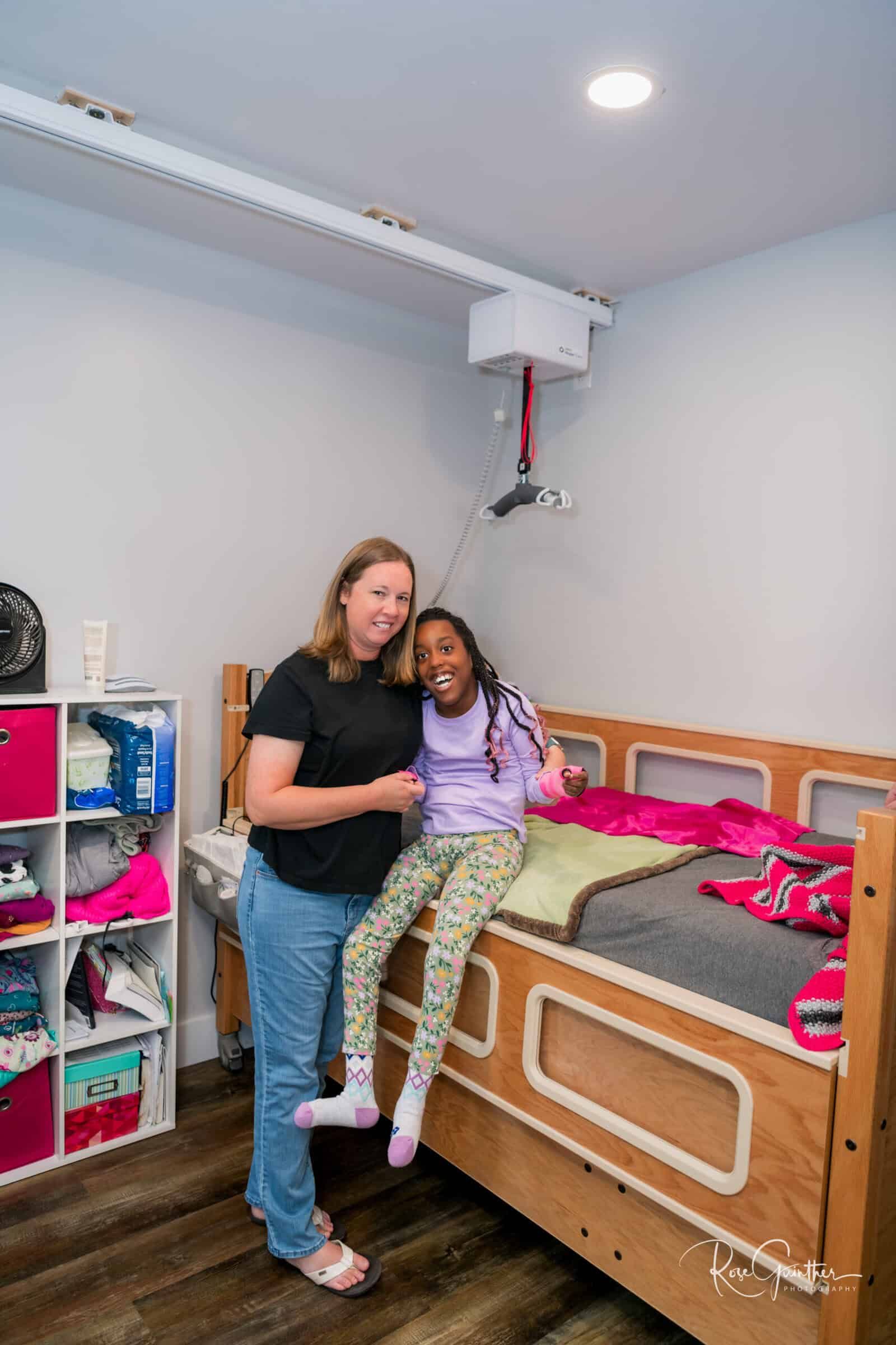 Woman and girl in accessible bedroom, smiling next to the bed. Ceiling lift installed above.
