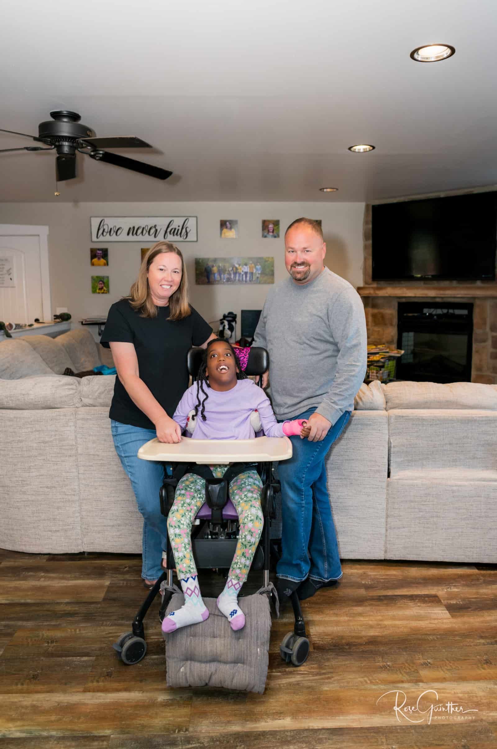 Family poses in living room; daughter in wheelchair, parents smile, neutral colors, sunny.