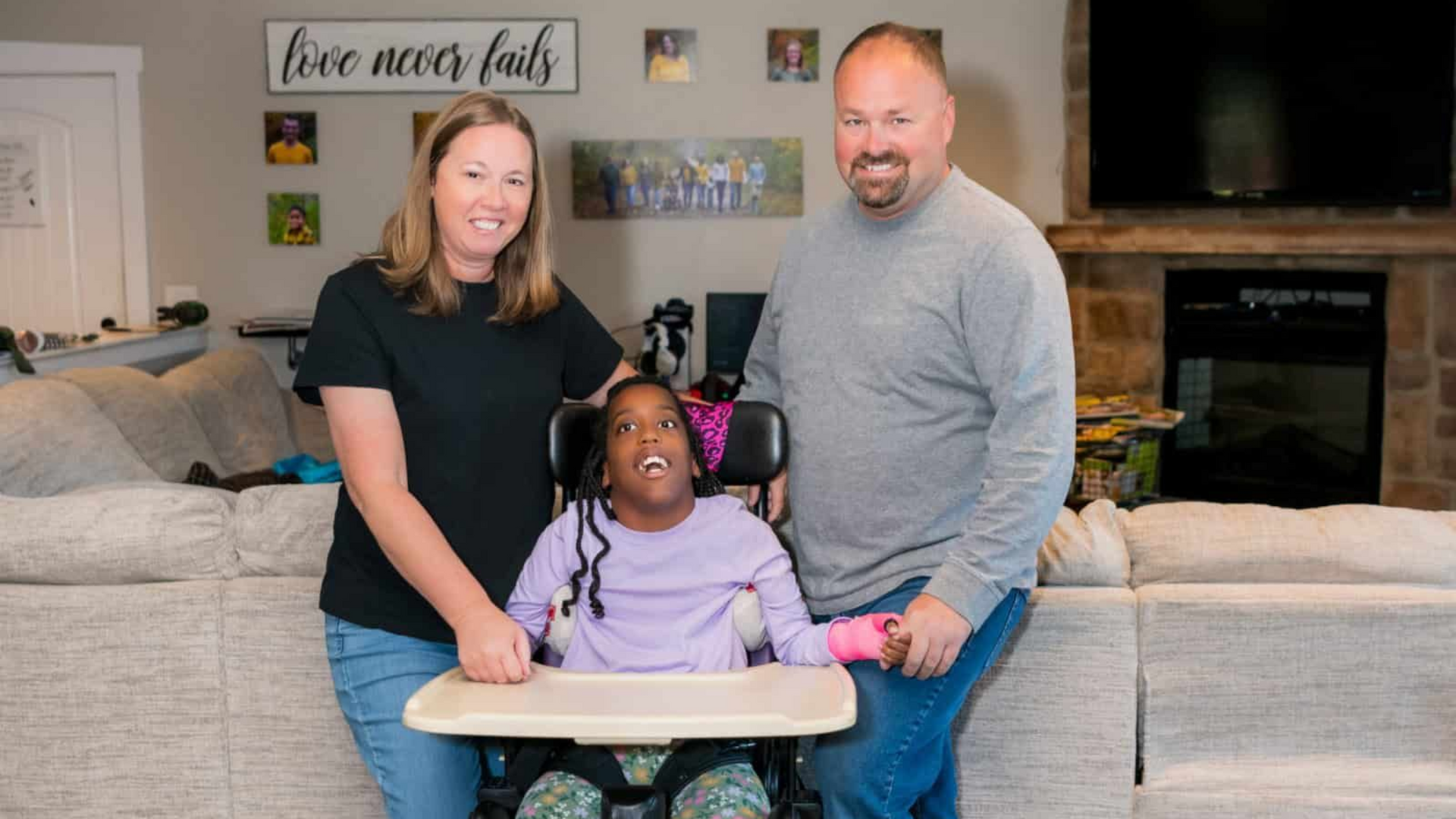 Family poses in living room; daughter in wheelchair, parents smile, neutral colors, sunny.