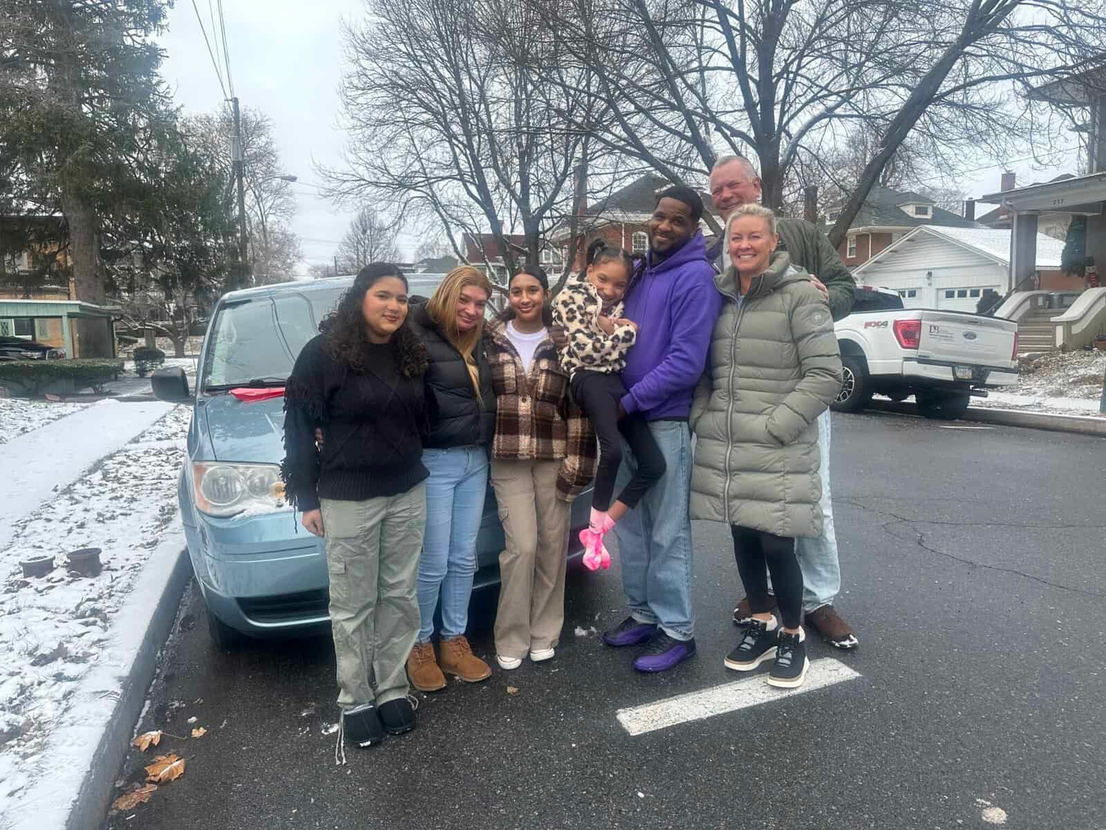 Group of people standing by a blue car on a snowy street, smiling.