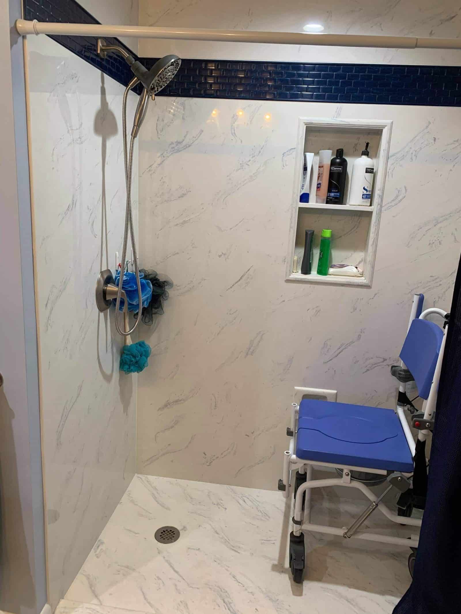 A wheelchair accessible shower with blue seat, marble-look walls, built-in shelf with products.