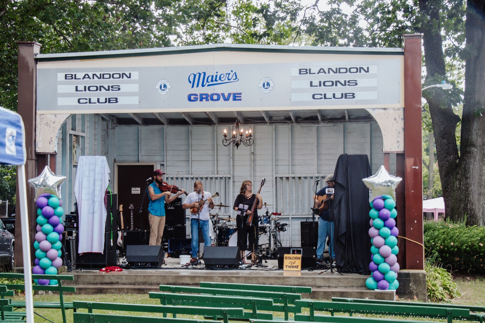 Group of six people smiling, posing under a blue canopy outdoors in a park setting.