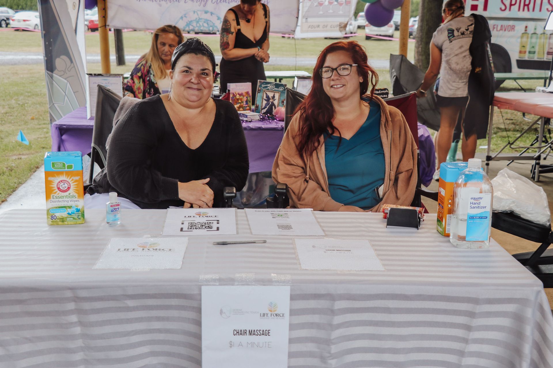 Two women sit at a table with signs. One in black shirt, other in blue. Outdoors, event setting.